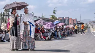 Two women in long dresses and wearing shawls stand under an umbrella - behind them a long line of voters, many under umbrellas, sit on a pavement by a tarmac road in Addis Ababa - 1 June 2026